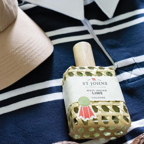 St Johns West Indian Lime cologne bottle in fishpot weave, styled on a navy striped shirt beside a beige cap.